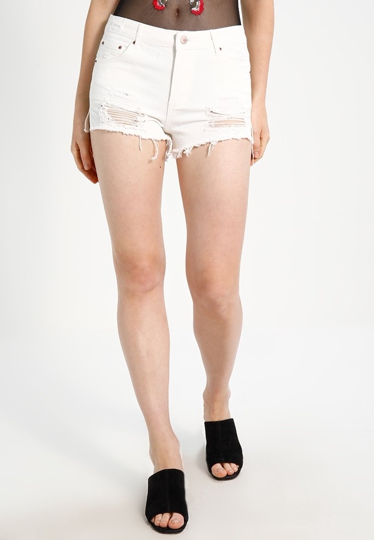 Mujer vistiendo shorts de mezclilla blanca rasgados, top de malla negra con bordado y tacones mule negros destalonados sobre un fondo blanco plano.