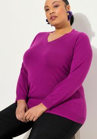 V-Ausschnitt lila Pullover aus weichem Strick mit langen Ärmeln, gerippten Bündchen und einer lockeren Passform. Über schwarzen Hosen getragen.