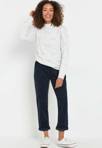 Mujer sonriente con cabello rizado, vistiendo un suéter de punto blanco, pantalones azul marino con puños y zapatillas blancas, de pie contra un fondo blanco.