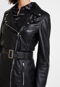 Svart läderbikerjacka med en figurnära design, asymmetrisk dragkedja, silverdetaljer, bältad midja och skjortkrage över en mönstrad topp.