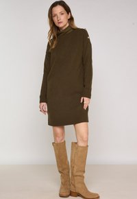 Abito lavorato a maglia verde oliva con scollatura alta e polsini a coste. Presenta dettagli con bottoni sulle spalle e un design rilassato, di lunghezza al ginocchio.