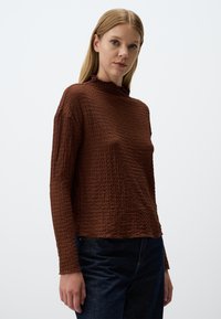 Haut à manches longues marron avec un tissu texturé, un col montant et une coupe décontractée. Présente un ourlet légèrement effiloché et un détail lisse au niveau des bras.
