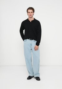 Czarna koszulka polo z długim rękawem i kołnierzykiem, zestawiona z jasnoniebieskimi luźnymi jeansami i czarnymi butami. Posiada kieszeń na piersi.