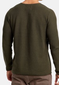 Mann in olivgrünem, gestricktem Langarmshirt und braunen Hosen, von hinten vor einem schlichten Hintergrund gezeigt.