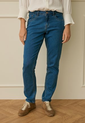 Personne portant un jean bleu, un chemisier crème avec des poignets froncés et des baskets en daim marron, debout sur un parquet en bois.