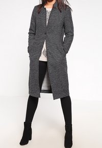 Cappotto bianco e nero a pois, con taglio dritto, maniche lunghe, tasche frontali e chiusura con un solo bottone, indossato sopra un maglione leggero.
