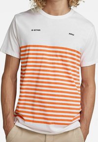 Hvid bomulds t-shirt med orange og hvide vandrette striber, prydet med sorte "G-STAR" og "RAW" logoer over det stribede område.