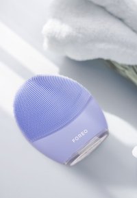 Spazzola per il viso Foreo in silicone viola chiaro, forma ovale con setole testurizzate, presenta una base liscia e il marchio nella parte anteriore.