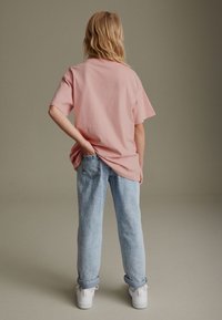 T-shirt di cotone rosa con maniche corte, vestibilità comoda, abbinata a jeans azzurri arrotolati e sneakers bianche, vista da dietro.
