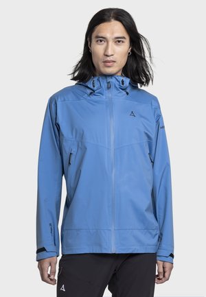 Person mit langen schwarzen Haaren, die eine blaue Kapuzenjacke mit Reißverschluss und schwarze Hose trägt, steht vor einem einfarbigen hellen Hintergrund.