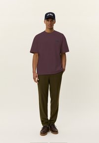 Bordo spalvos trumpomis rankovėmis marškinėliai su subtiliu logotipu, derinami su alyvuogių žalių spalvų kelnėmis ir rudi batai su raišteliais, papildyti tamsiai mėlyna kepure.