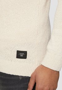 Crème-kleurige gebreide trui met een gestructureerd oppervlak, geribde manchetten en een zwart label met witte tekst aan de onderkant.