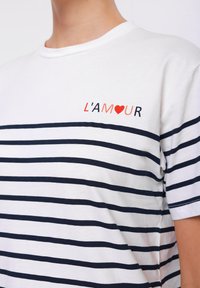 T-shirt en coton blanc avec des rayures horizontales marines, présentant un petit graphic L'amour rouge et noir sur la poitrine. Design classique à col rond.