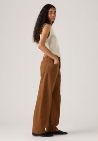 Vrouw met lang krullend haar, gekleed in een mouwloos wit gehaakt topje, brede bruine broek en zwarte schoenen, staande met handen in de zakken.