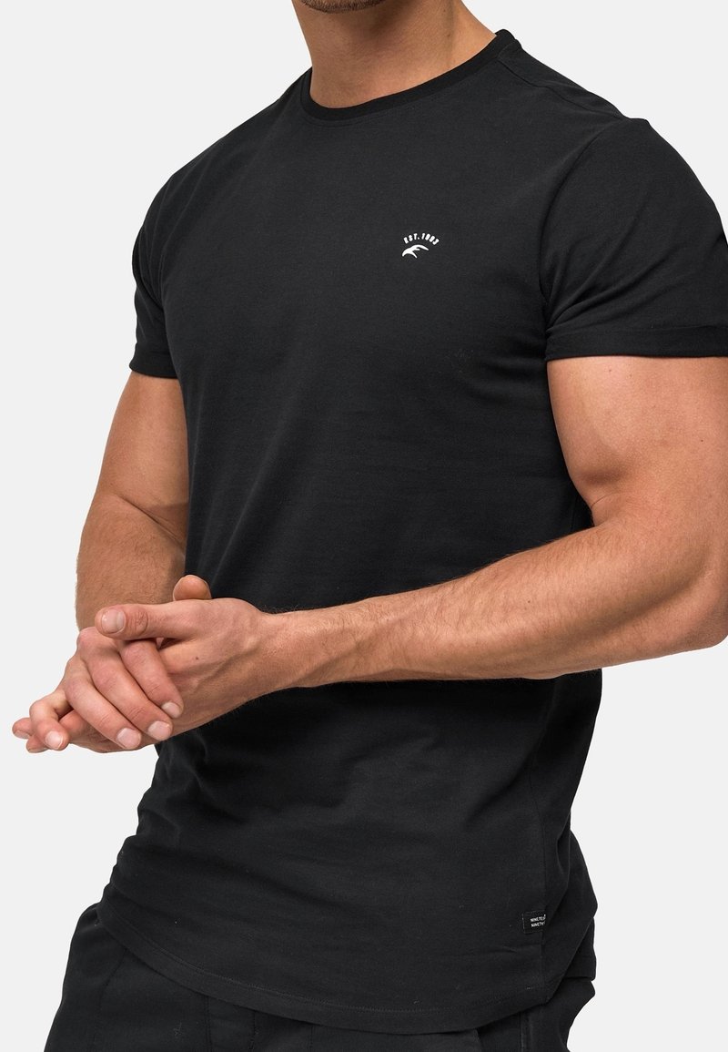 Homme aux bras musclés portant un t-shirt noir ajusté avec un petit logo blanc sur la poitrine gauche et les mains jointes.