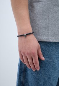 Mão a usar um bracelete de contas pretas com um encantamento de sol prateado, repousando ao lado de umas calças de ganga azuis e uma t-shirt cinza.