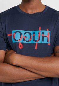 T-shirt blu navy in cotone, con un audace design grafico che presenta la scritta "HUGO" in colori blu e rosso al centro del petto. Maniche corte.