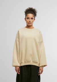 Felpa beige oversize con scollatura a girocollo a costine, maniche lunghe e un sottile logo bianco sul petto. Tessuto morbido e strutturato.