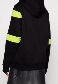 Sudadera negra con diseño oversize, que presenta franjas horizontales de un brillante color verde lima en las mangas y un detalle de cremallera en el frente. Textura suave.