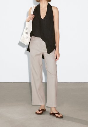 Femme portant un haut asymétrique noir sans manches, un pantalon beige à jambes larges, des sandales marron et tenant un sac en crochet blanc.