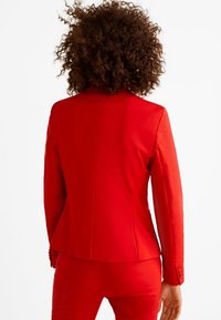 Rode op maat gemaakte blazer met een gestructureerde pasvorm, enkele zijsplit aan de achterkant en gestylde mouwen met knoopaccenten. Gladde, textuurrijke stof.