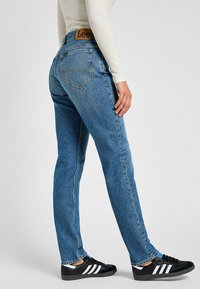 Jeans de mezclilla azul claro con corte recto, que cuentan con una etiqueta de cuero color marrón, diseño de cinco bolsillos y un sutil desgastado en la tela.