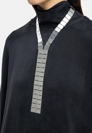 Osoba ubrana w czarny wysokookryty kołnierz z długim srebrnym naszyjnikiem z segmentów z metalu wiszącym na szyi i piersi.