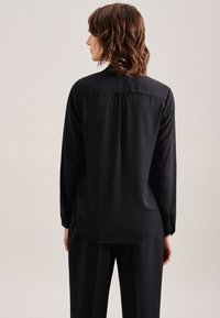 Langärmlige schwarze Bluse, glattes Material, lässige Passform, abgerundeter Saum, Rückenfalten-Detail, mit Knöpfen an den Manschetten, getragen mit passenden schwarzen Hosen.