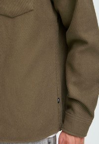 Olive green ribbed long-sleeve shirt with a chest pocket, side slits, and visible stitching detail on the hem and cuffs. Soft texture.