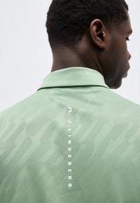 Άνδρας με κοντά σγουρά μαλλιά, που φοράει ένα ανοιχτό πράσινο πουκάμισο με υφή και έναν κάθετο λογότυπο "J.LINDEBERG" στο πίσω μέρος του γιακά.