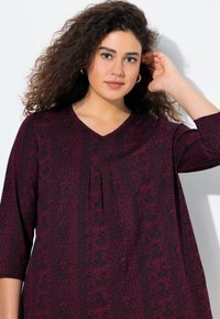 Haut maroon à motifs avec un décolleté en V et des manches ¾. Le tissu présente un design floral texturé, créant un look moderne et décontracté.