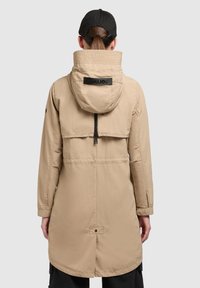 Parka beige con capucha, que presenta un cuello alto, cierre de cremallera, puños elásticos y una abertura trasera para transpirabilidad. Marca KHUJO exhibida.