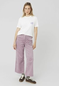 T-shirt blanche avec un graphique violet, associée à un pantalon large en velours côtelé violet clair. Des baskets marron foncé et des chaussettes à motifs sont visibles.