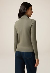 Maglione aderente verde oliva con colletto alto e maniche lunghe, dotato di dettagli con bottoni sui polsini. Il materiale sembra morbido con una superficie liscia.