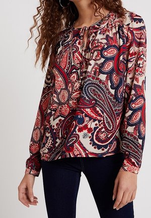 Femme portant un chemisier à manches longues avec des motifs paisley rouges, bleu marine et beige, associé à un pantalon bleu foncé.