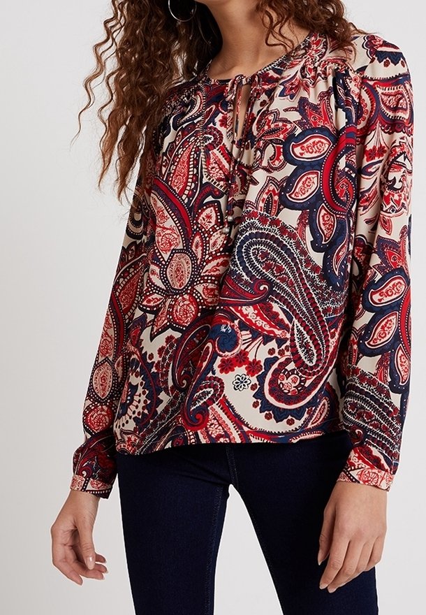 Femme portant un chemisier à manches longues avec des motifs paisley rouges, bleu marine et beige, associé à un pantalon bleu foncé.