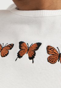 White ribbed fabric featuring three embroidered orange butterflies with black accents, positioned near the neckline.