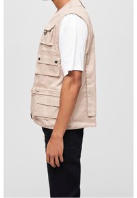 Beige tygväst med flera framfickor, knappdetaljer och en avslappnad passform. Bärs över en vit t-shirt och kombineras med mörka byxor.