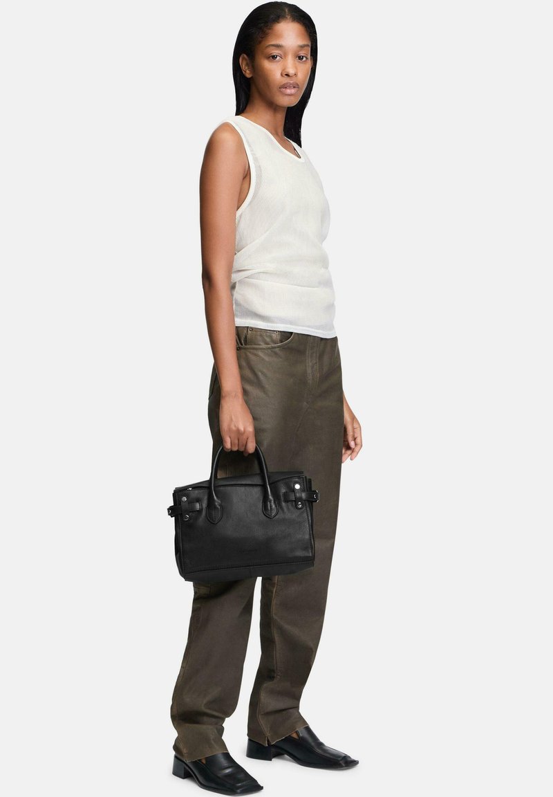 Sac à main en cuir noir avec des poignées doubles, présentant une forme structurée et des accents de quincaillerie subtils. Porté avec un débardeur blanc et un pantalon olive.
