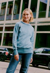 Felpa azzurro chiaro con motivo screziato, collo rotondo e dettagli con bottoni sulla spalla. Abbinata a jeans di denim scuro.