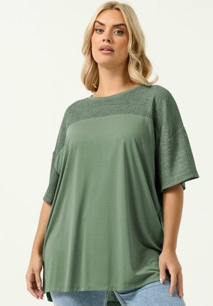 Lyshåret kvinde iført en løs, salviegrøn kortærmet top med struktureret overdel og ærmer, kombineret med lyseblå jeans.