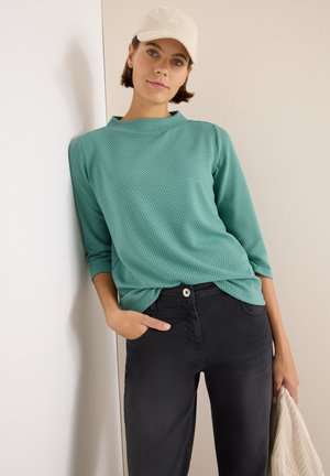 Mintgrünes, strukturiertes Langarmoberteil, kombiniert mit schwarzen Hosen. Das Model hält eine cremefarbene Tasche und trägt eine helle Kappe.
