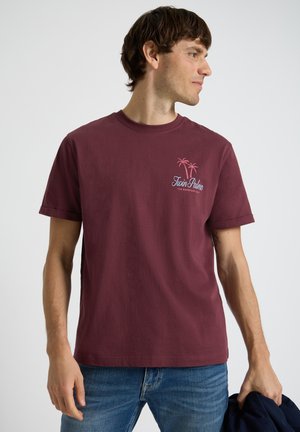 Mężczyzna w bordowej koszulce "Twin Palms" i niebieskich dżinsach, trzyma ciemną kurtkę, patrzy w prawo na białym tle.