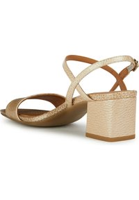 Sandales à talons beiges avec un dessus en cuir texturé, bout ouvert et lanières croisées. Talon carré, semelle intérieure lisse et bride de cheville réglable.