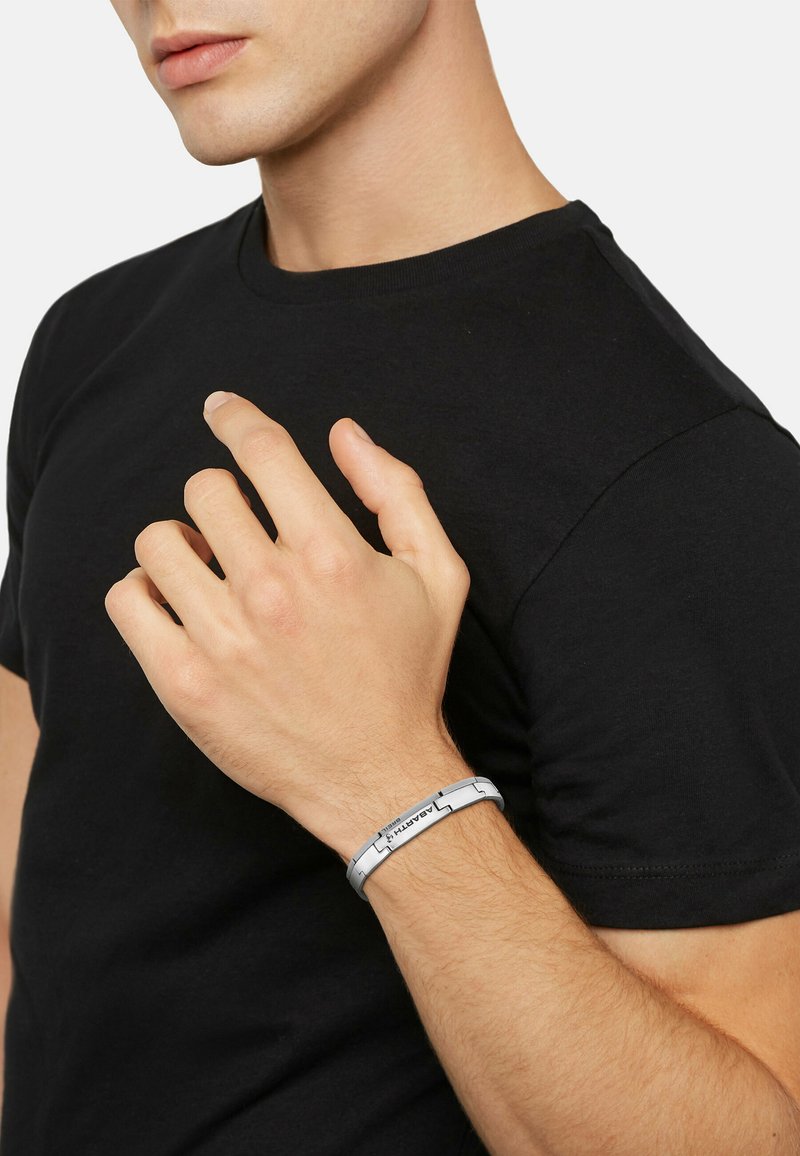 Silverfärgad armband med sammanlänkade metallkedjor, som har en slät yta och ingraverade detaljer. Bärs på handleden mot en svart skjorta.