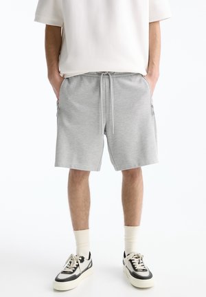 Homme portant un short gris clair à cordon, des chaussettes blanches, des baskets noir et blanc, et une chemise blanche à manches courtes, debout les mains dans les poches.