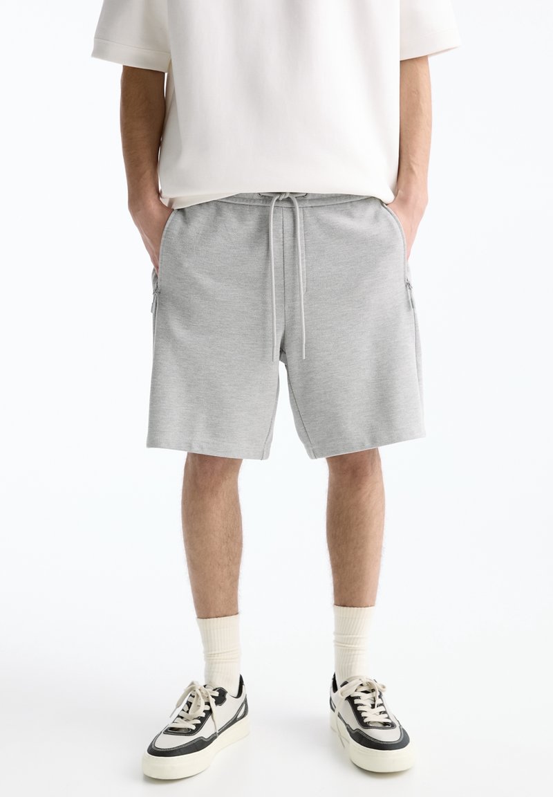 Homme portant un short gris clair à cordon, des chaussettes blanches, des baskets noir et blanc, et une chemise blanche à manches courtes, debout les mains dans les poches.