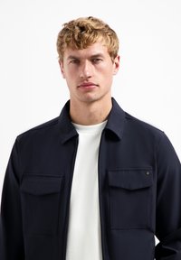 Marineblauwe ritsjack met een kraag, twee borstzakken en een gladde stofstructuur, gedragen over een wit shirt.