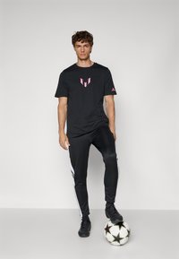 Sort atletisk T-shirt med pink logo, parret med sorte joggers med hvide sidelister, stående på en fodbold.