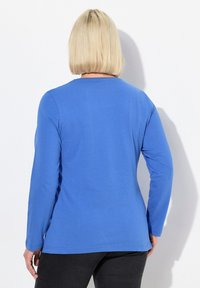 Langärmliges blaues T-Shirt aus Baumwolle, schlanker Schnitt, runder Halsausschnitt, glatte Textur, mit geradem Saum. Getragen mit schwarzen Hosen.